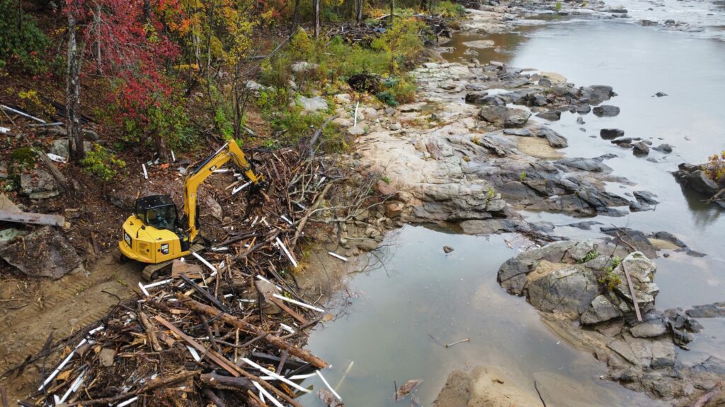 Excavator operates onshore to pick up debris from a large pile